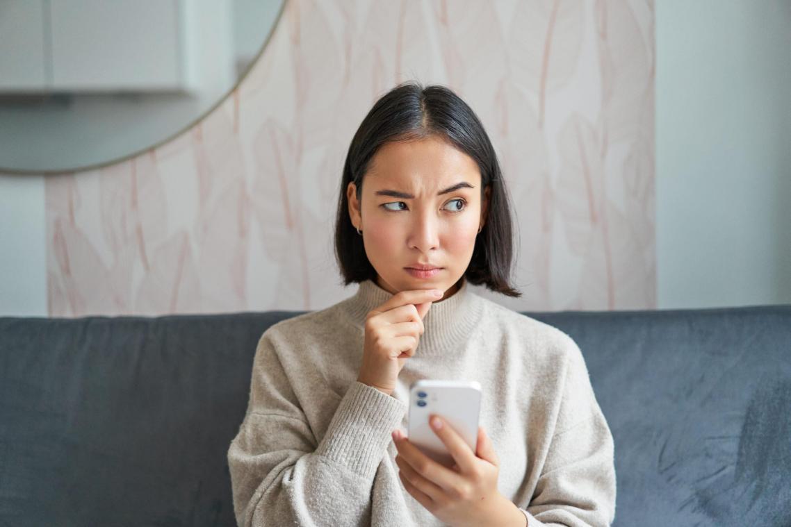 Portrait of girl sitting on sofa with smartphone, looking thoughtful and hesitant at mobile phone screen.